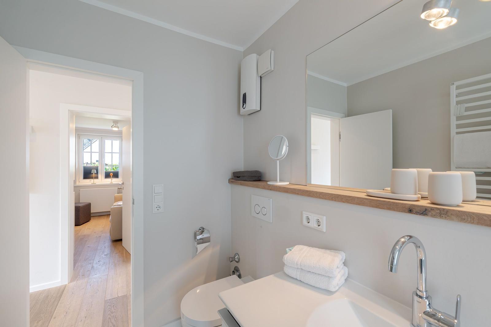 Badezimmer mit Waschbecken, Spiegel und Toilette. Durchgang zu Schlafzimmer mit Holzboden.