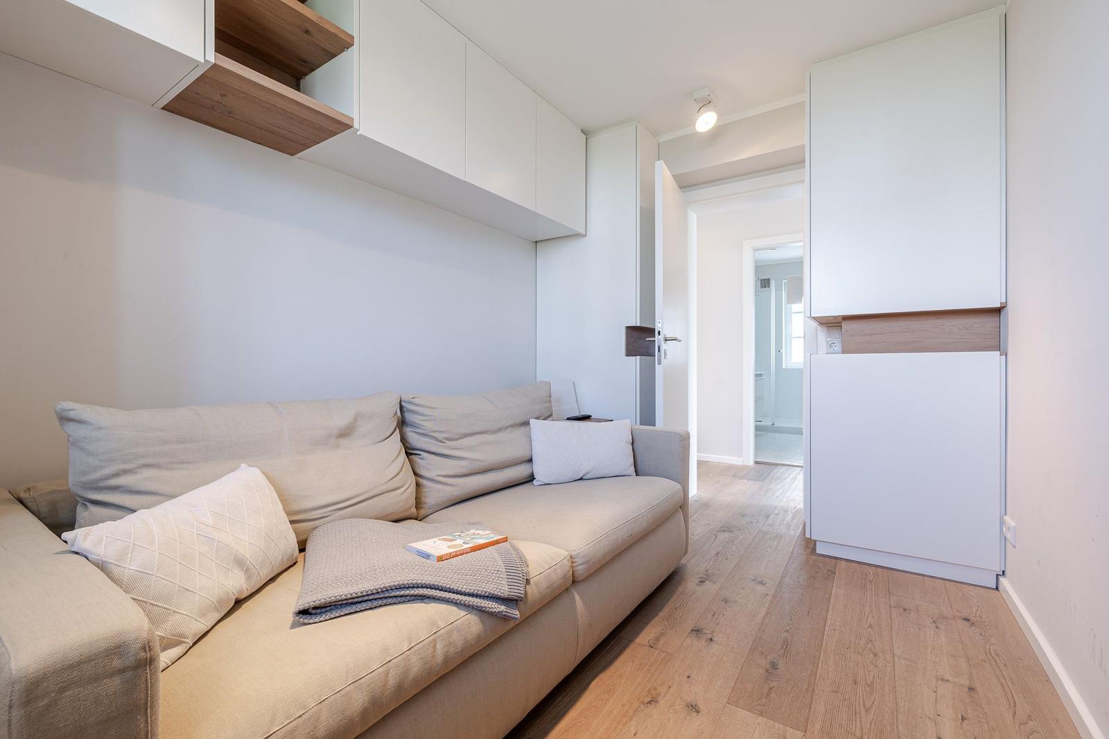 Wohnzimmer mit Sofa, Holzboden und weißen Schränken. Bücher und Decke liegen auf dem Sofa.