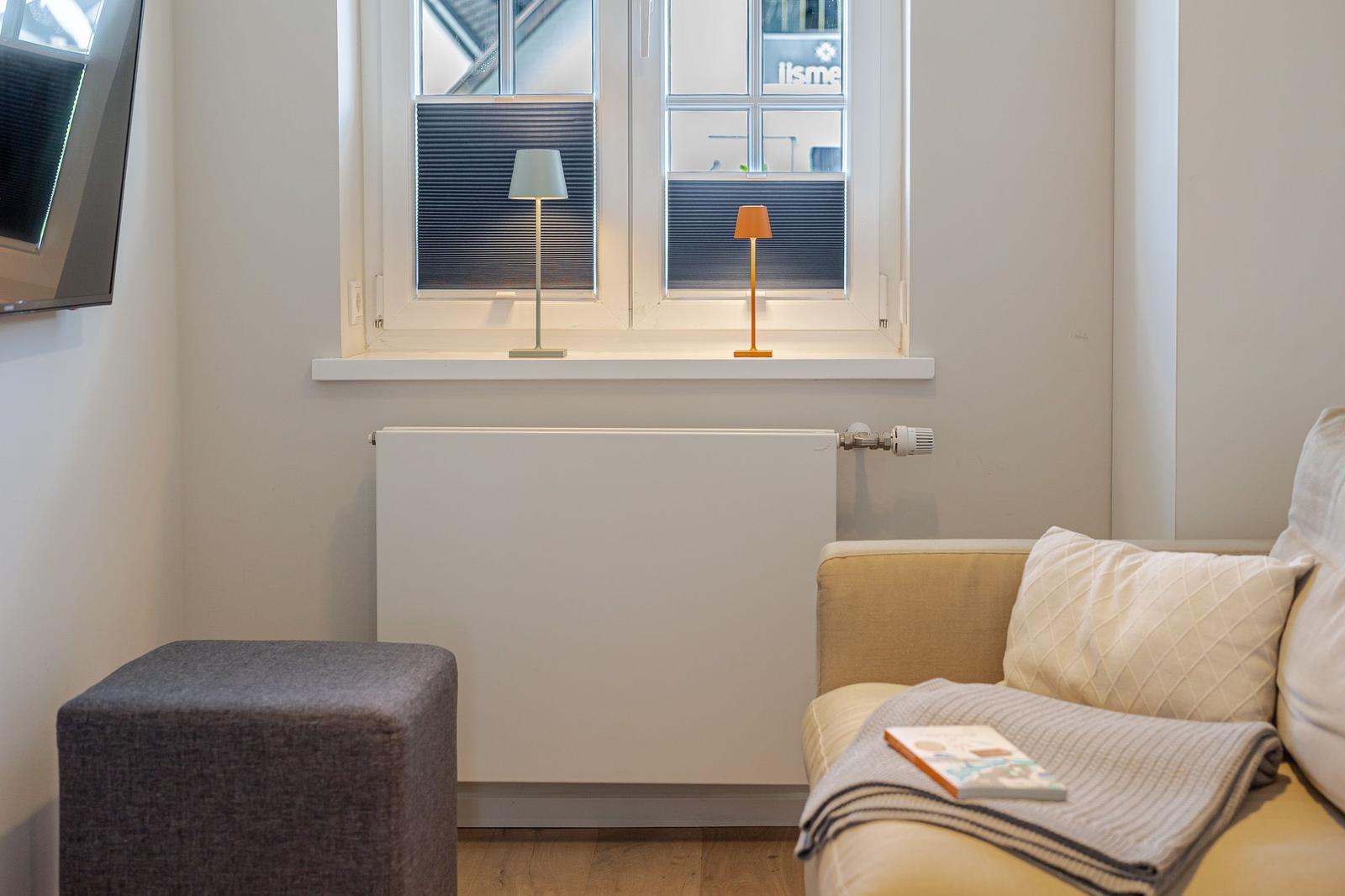 Ein Wohnbereich mit Sofa, Fußhocker und zwei Lampen am Fenster.