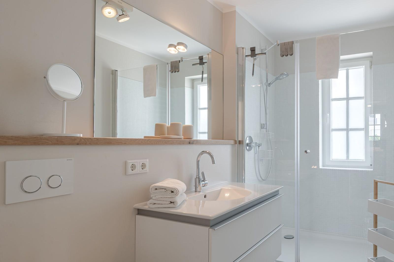 Badezimmer mit Dusche, Waschbecken und Spiegel. Fenster sorgt für Tageslicht.