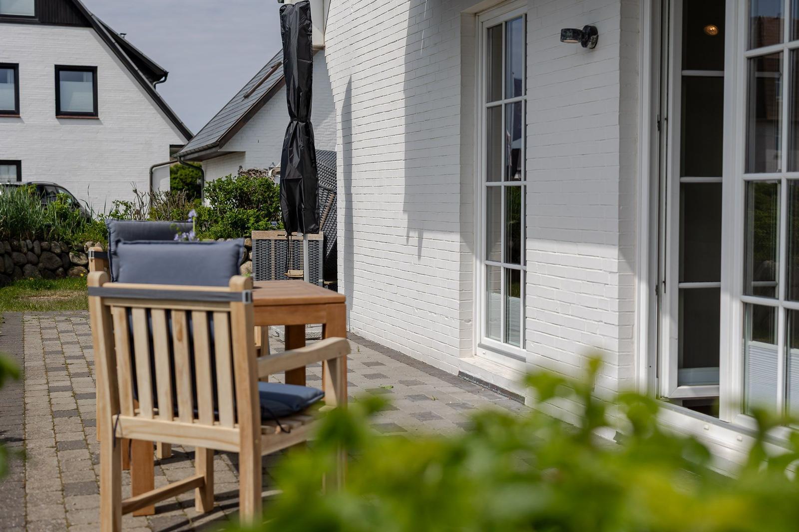 Terrasse mit Holztisch und Stühlen vor weißer Hauswand mit Fenstern.