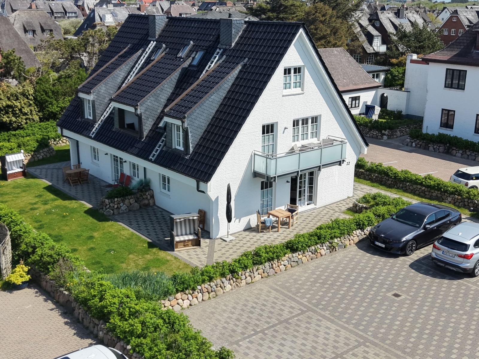Weißes Haus mit dunklem Dach, Terrasse und Garten. Parkplatz mit zwei Autos.