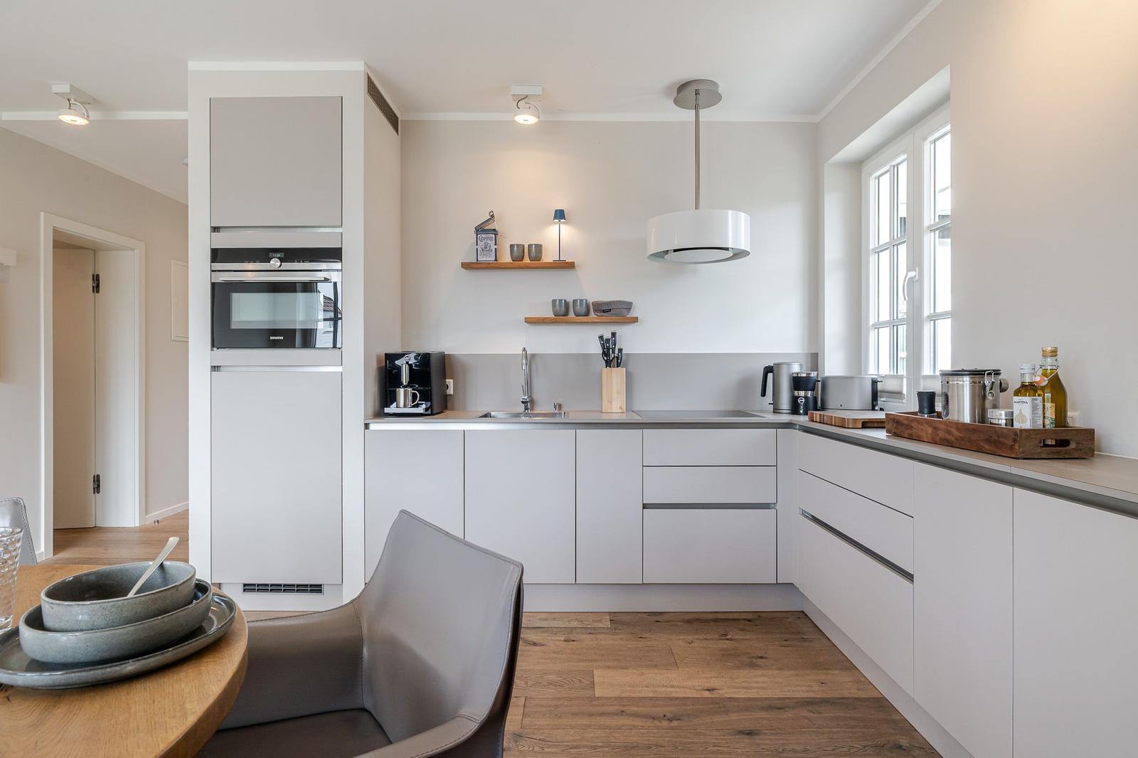 Moderne Küche mit weißer Einbauküche, Edelstahlspüle und Holzboden. Fenster und Deckenleuchten sorgen für Licht.