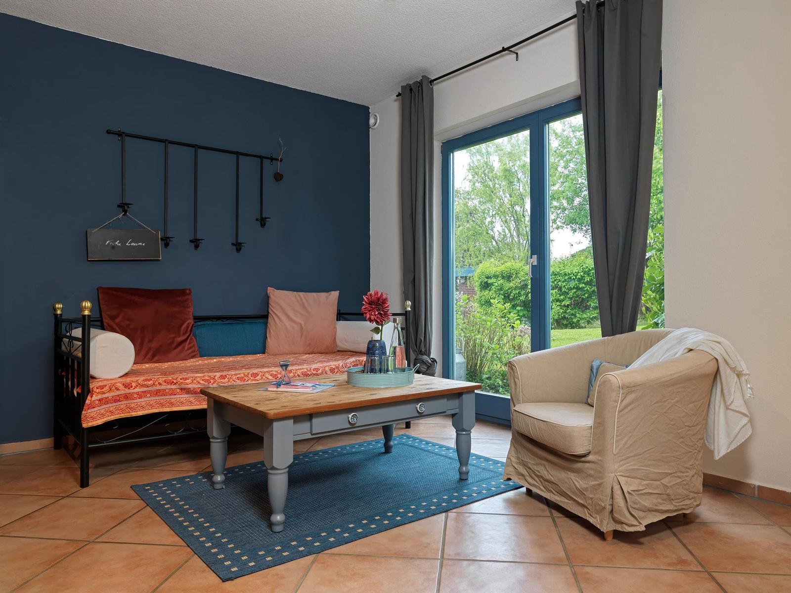 Ein Raum mit Bett, Couch und Tisch. Durch Fenster blickt man auf Garten.