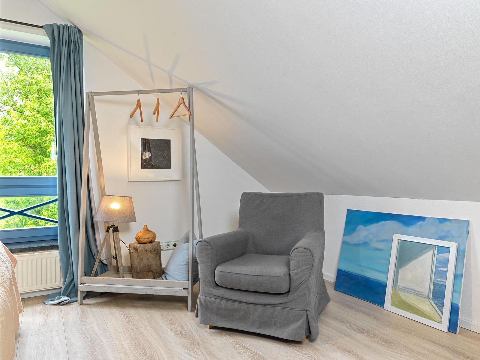 Zimmer mit grauem Sessel, Kleiderständer und Fenster mit Blick auf Grün.