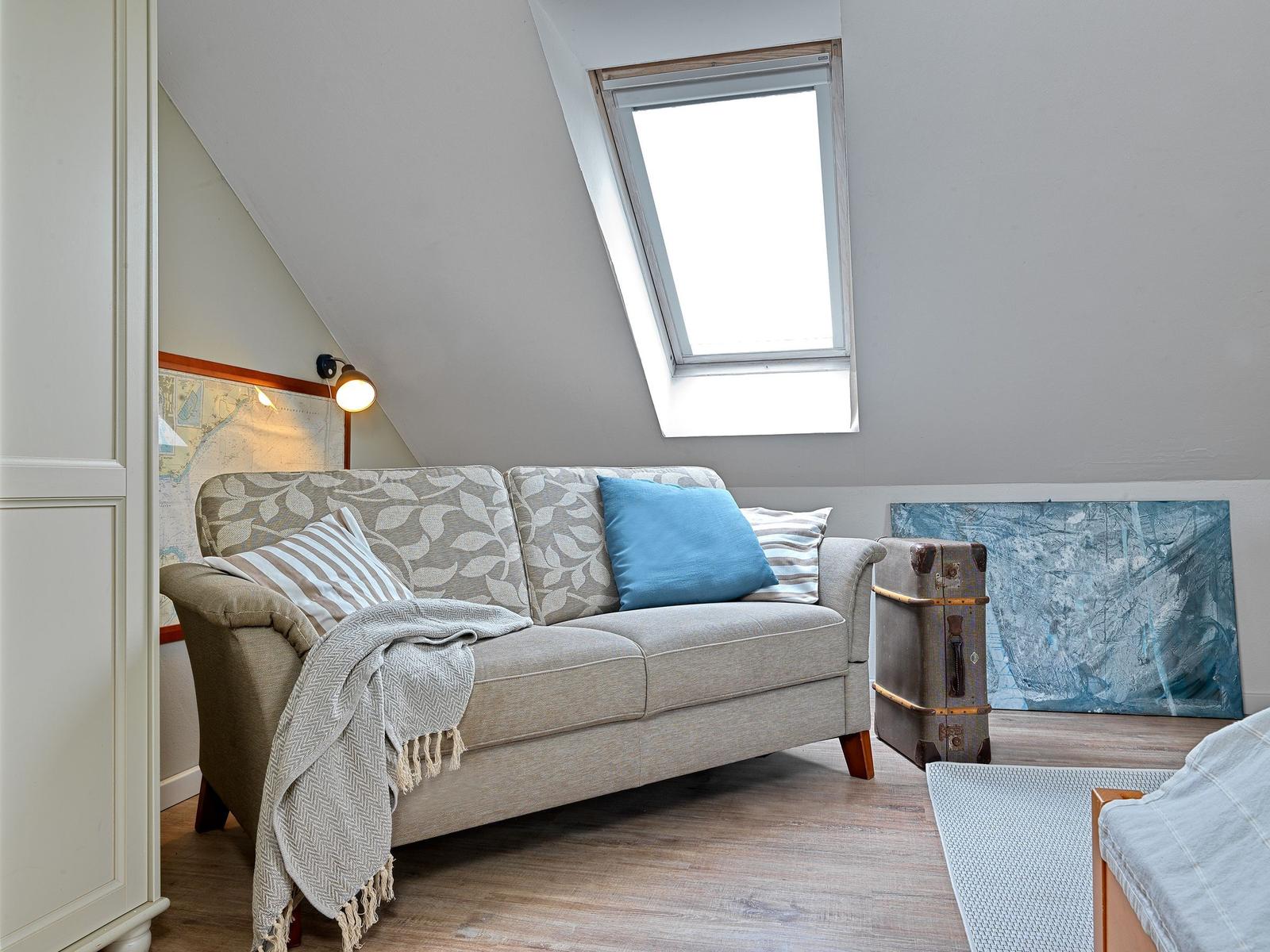 Ein Sofa mit Kissen und Decke unter einem Dachfenster. Ein Koffer und ein Bild sind sichtbar.
