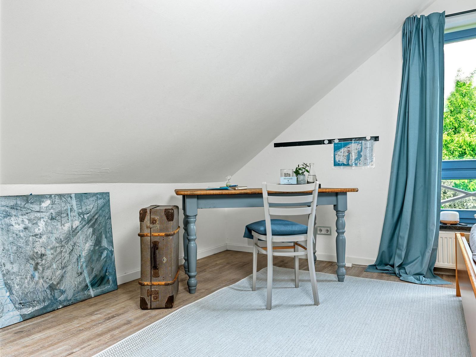 Schlafzimmer mit Schreibtisch, Stuhl und Koffer. Fenster mit Vorhang und Wandbild.