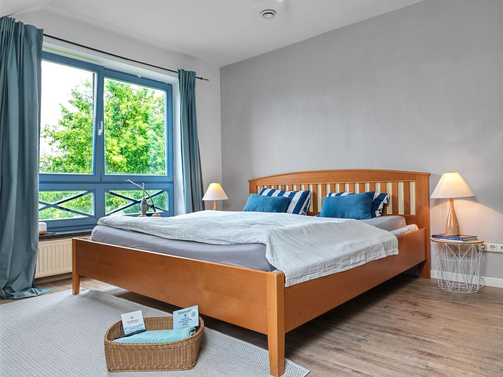 Schlafzimmer mit Holzbett, blauen Kissen und großem Fenster mit Blick auf Bäume.