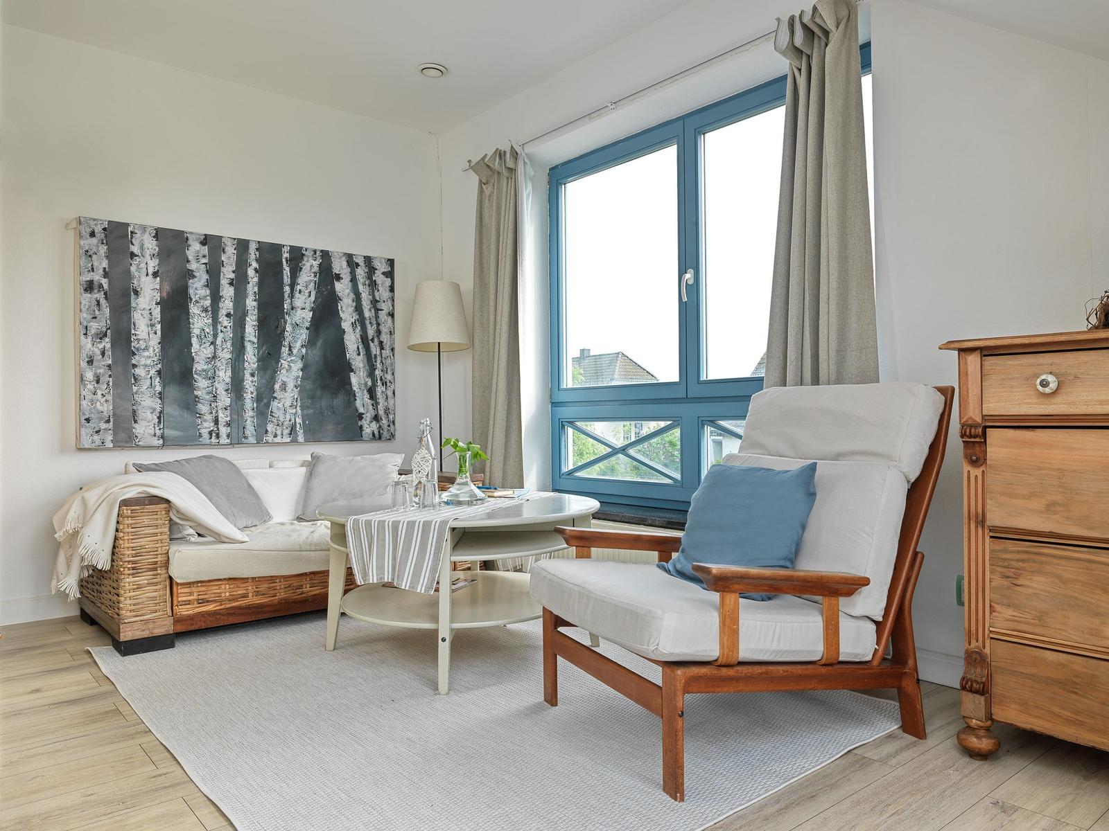 Wohnzimmer mit Sofa, Sessel, Tisch und Fenster mit blauem Rahmen.