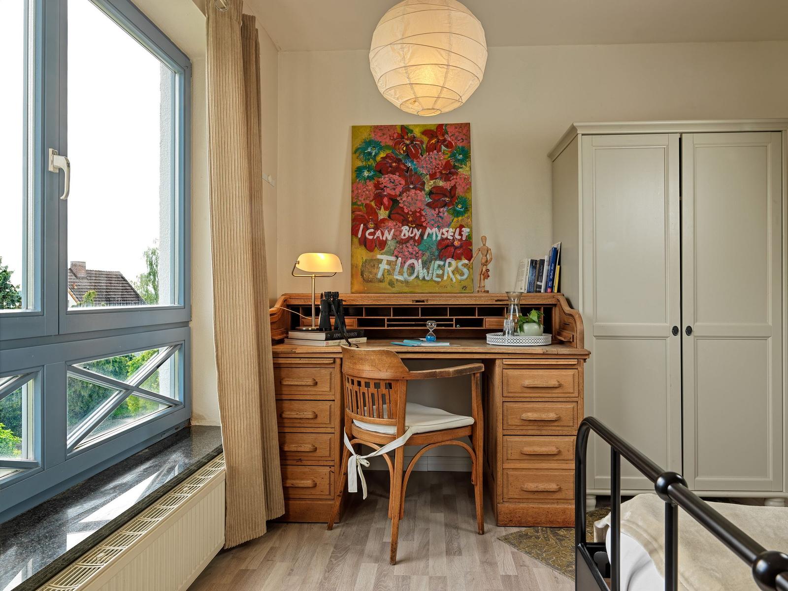 Schreibtisch mit Stuhl, Wandbild und Kleiderschrank neben Fenster mit Blick nach draußen.