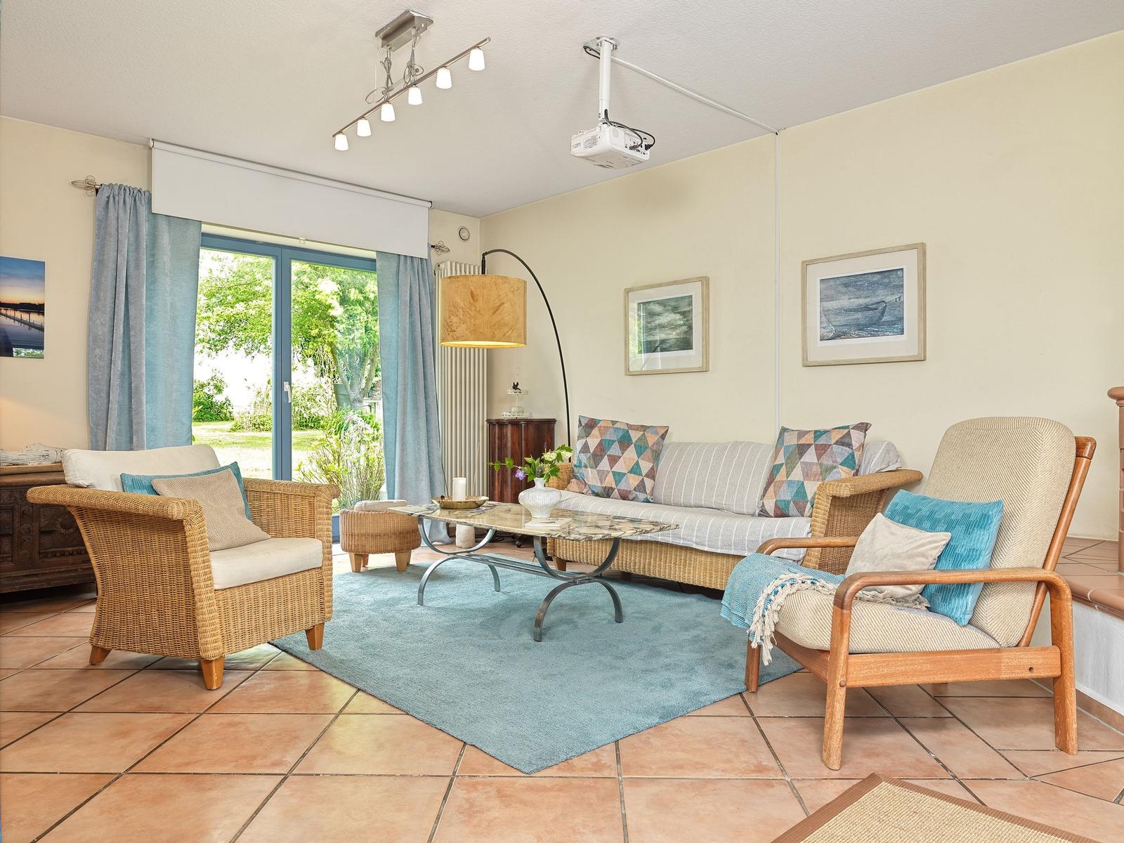 Wohnzimmer mit Sofa, Sesseln und Glas-Tisch. Fenster mit Blick auf Garten. Projektionssystem an Decke.
