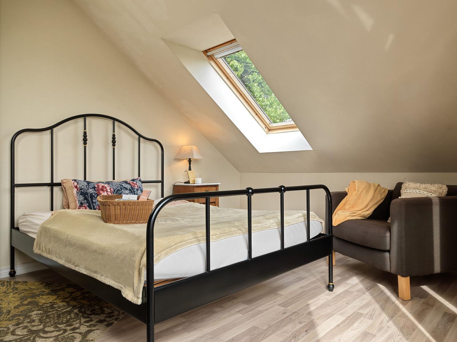 Schlafzimmer mit Dachschrägen, Bett, Sessel und Dachfenster mit Blick auf Grün.