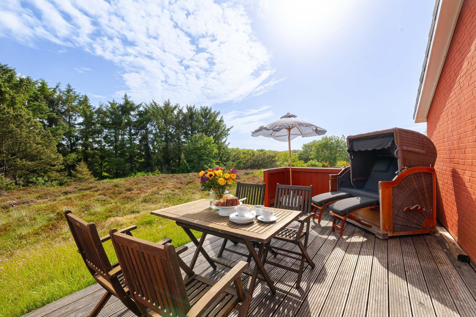 Terrasse mit Tisch, Stühlen, Sonnenschirm und Strandkörbe. Blick auf grüne Wiesen und Bäume.