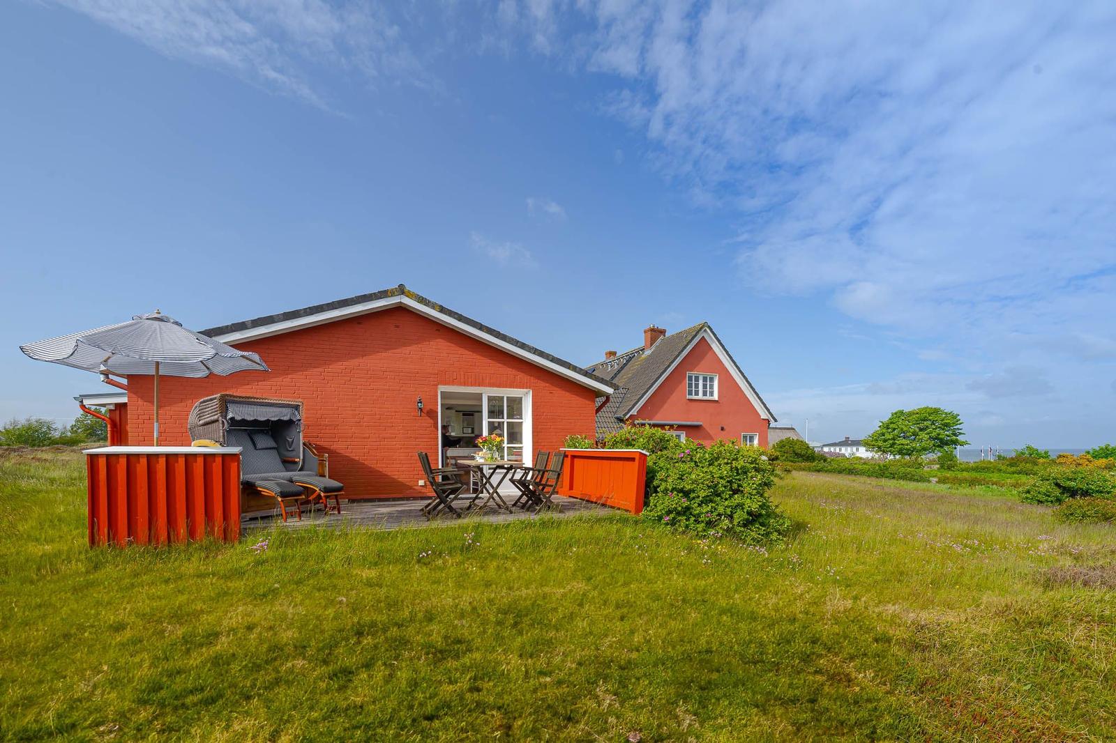 Rotes Haus mit Terrasse, Garten und Sonnenschirm. Blick auf grünes Feld und Himmel.