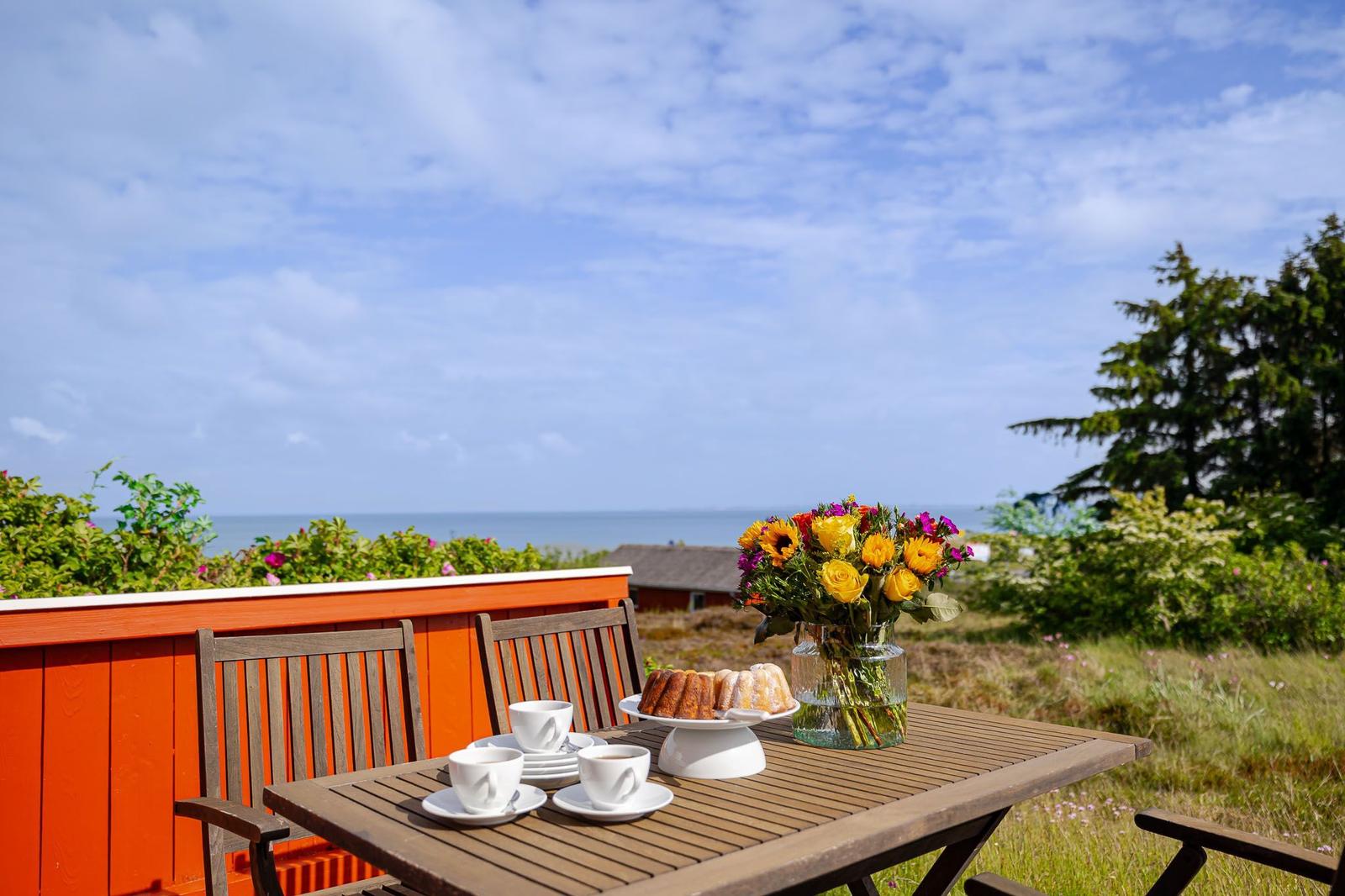 Terrasse mit Tisch, Stühlen, Blumen und Gebäck. Blick auf Meer und Himmel.