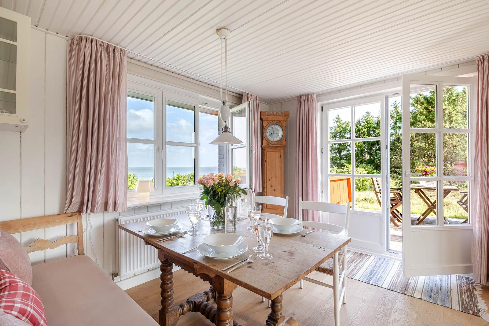 Esszimmer mit Holztisch, Fenstern zum Meer und Terrasse. Holzdecke, rosa Vorhänge.