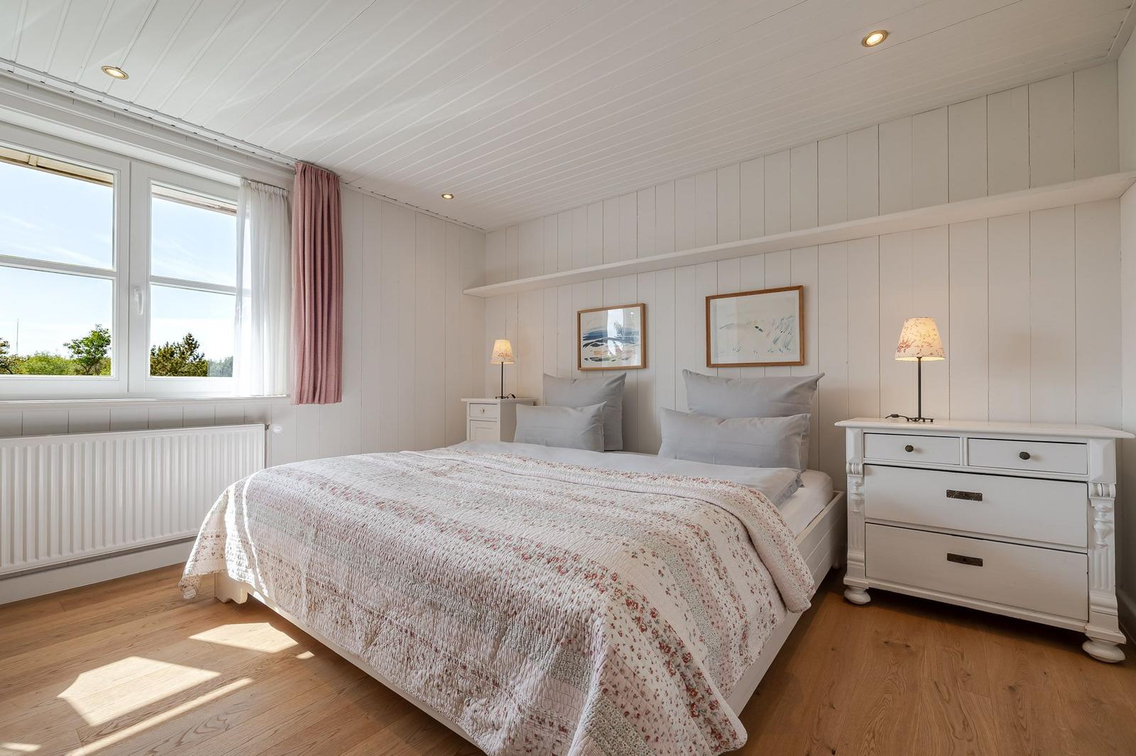 Schlafzimmer mit Doppelbett, weißen Wänden und Holzboden. Fenster mit rosa Vorhang.