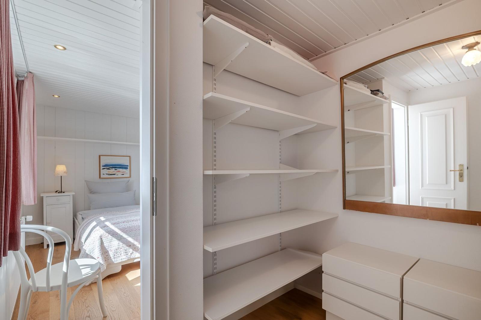 Schrank mit weißen Regalen und Spiegel. Blick in Schlafzimmer mit Bett und Nachttisch.