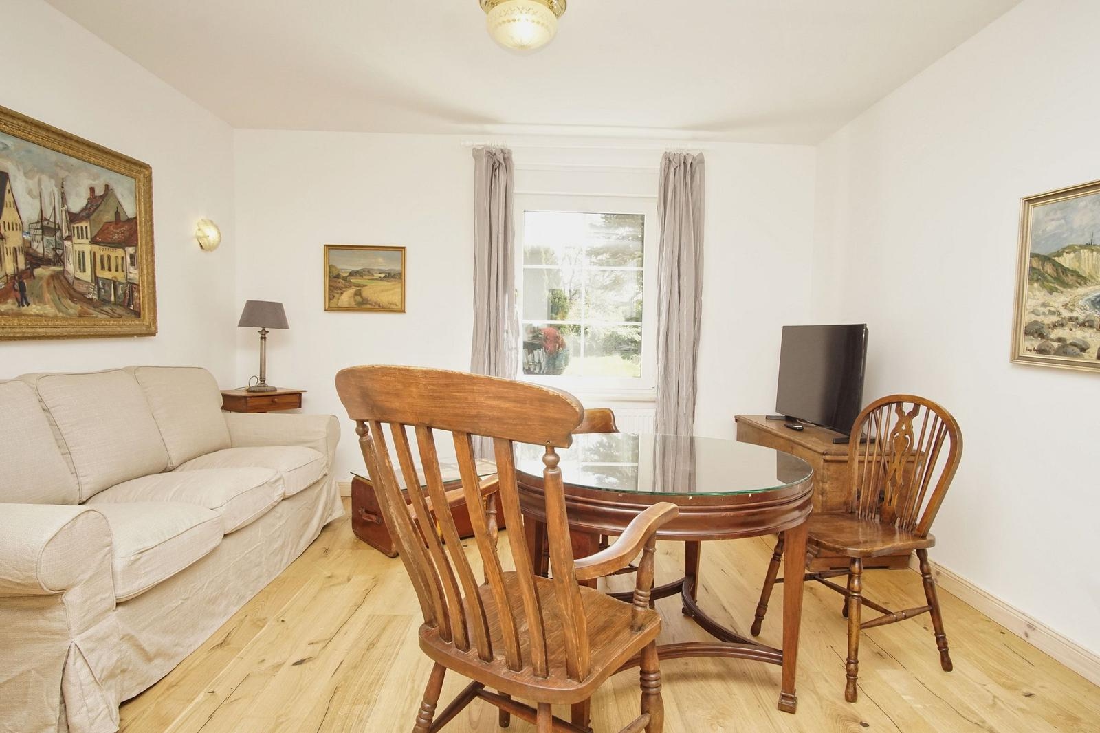 Wohnzimmer mit Sofa, Esstisch und Fernseher. Holzfußboden und Fenster mit Vorhängen.