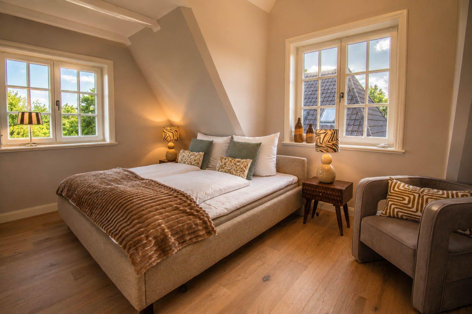 Schlafzimmer mit Doppelbett, zwei Fenstern und Sessel. Holzboden und Deckenbalken sichtbar.