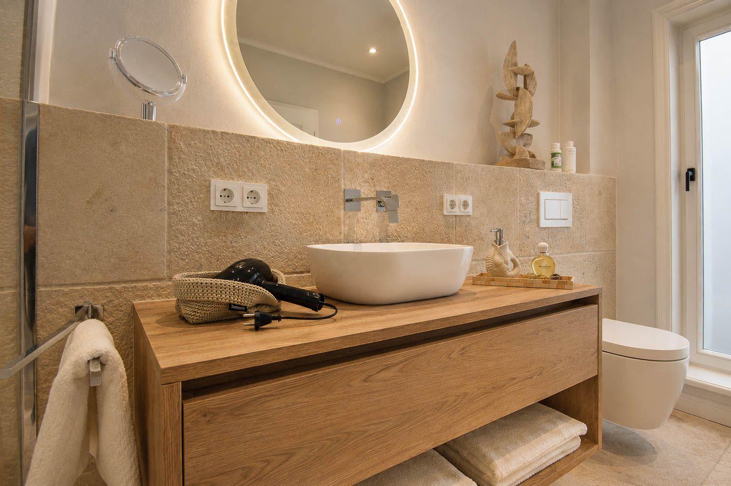 Bad mit Waschtisch, Spiegel und Toilette. Holzwaschtisch, Handtuchhalter und Fenster sichtbar.