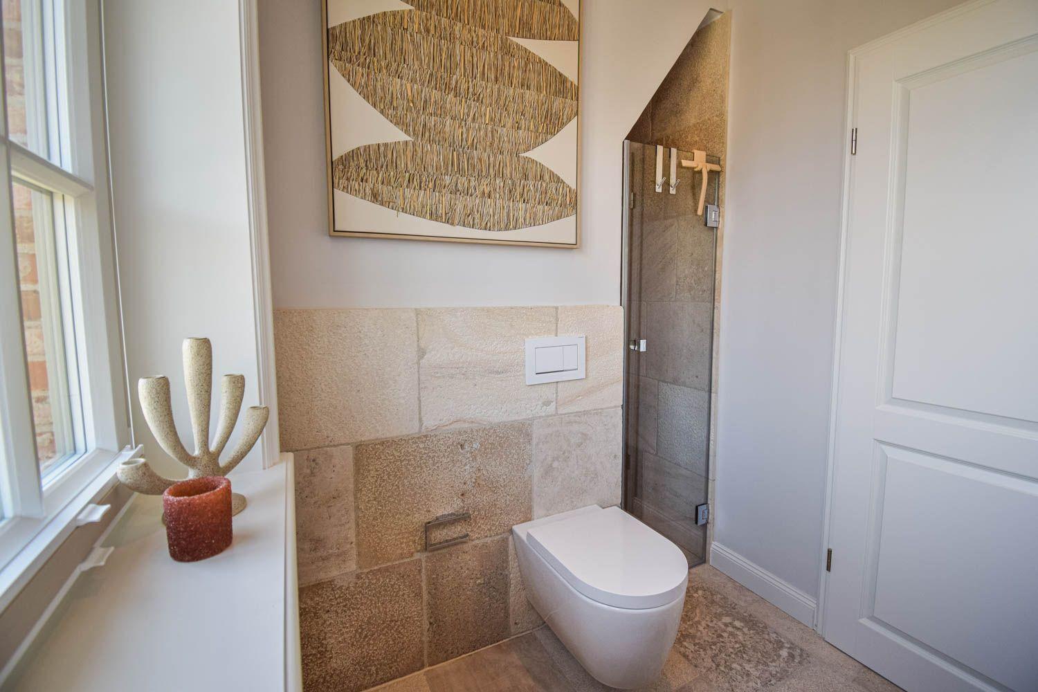 Badezimmer mit Toilette, Dusche und Fenster. Wandverkleidung aus Stein und Kunstwerk an der Wand.