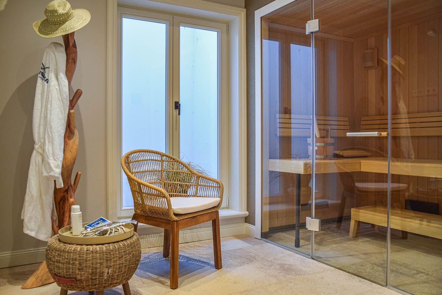 Sauna mit Glaswand, Stuhl und Hocker neben Fenster. Handtuch und Hut an Holzständer.