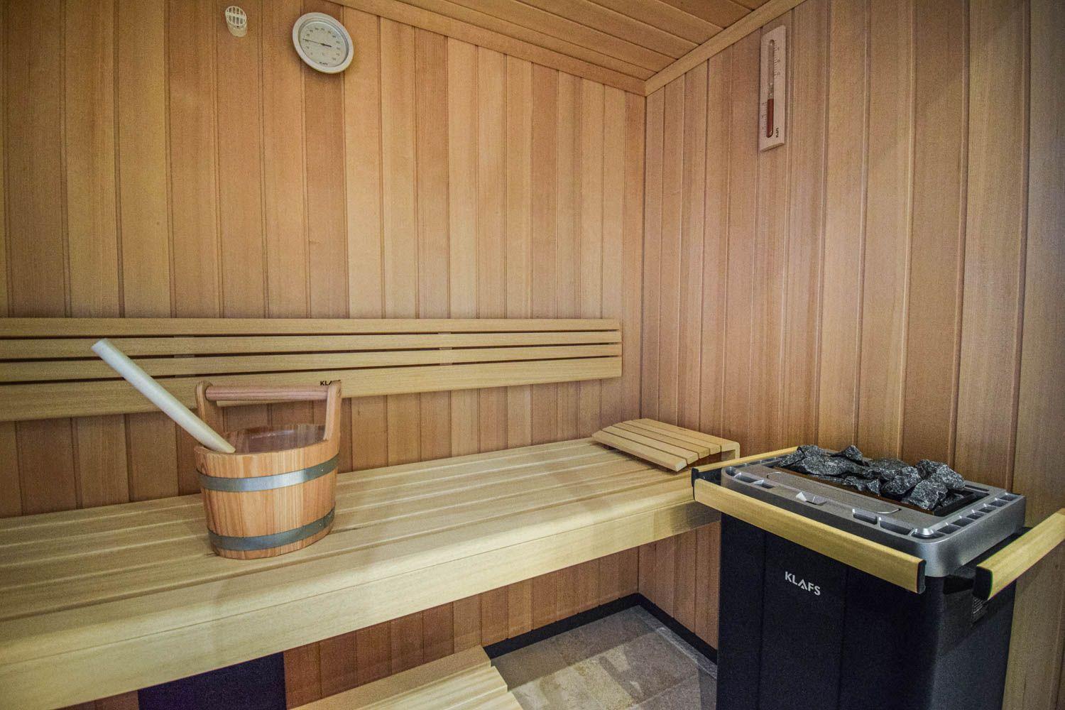 Sauna mit Holzbank, Eimer und Heizung. Wandthermometer und Holzwand.