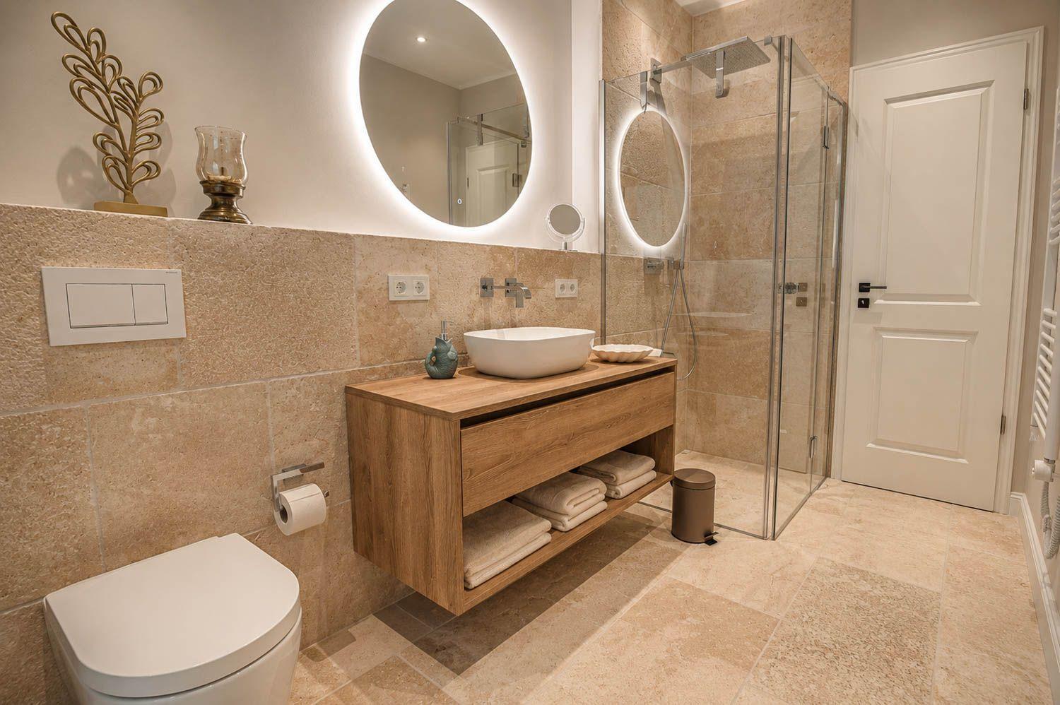 Badezimmer mit Dusche, Waschtisch und Toilette. Holzunterschrank und runder Spiegel.