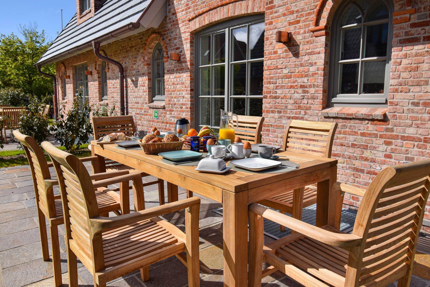 Holztisch mit Stühlen auf Terrasse vor Backsteinhaus mit Frühstückszubehör.