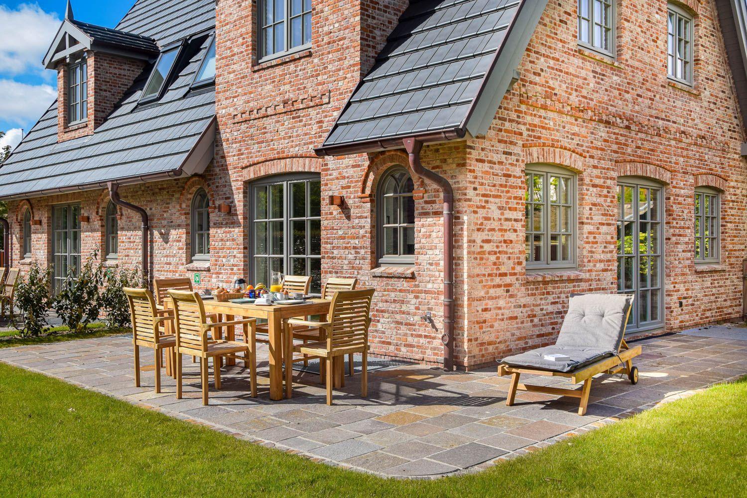 Terrasse mit Holztisch, Stühlen und Liegestuhl vor Backsteinhaus mit Dachgauben.
