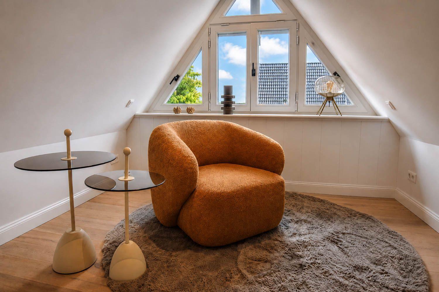 Ein Raum mit einem orangefarbenen Sessel, zwei Tischen und einem runden Teppich vor einem Dachfenster.