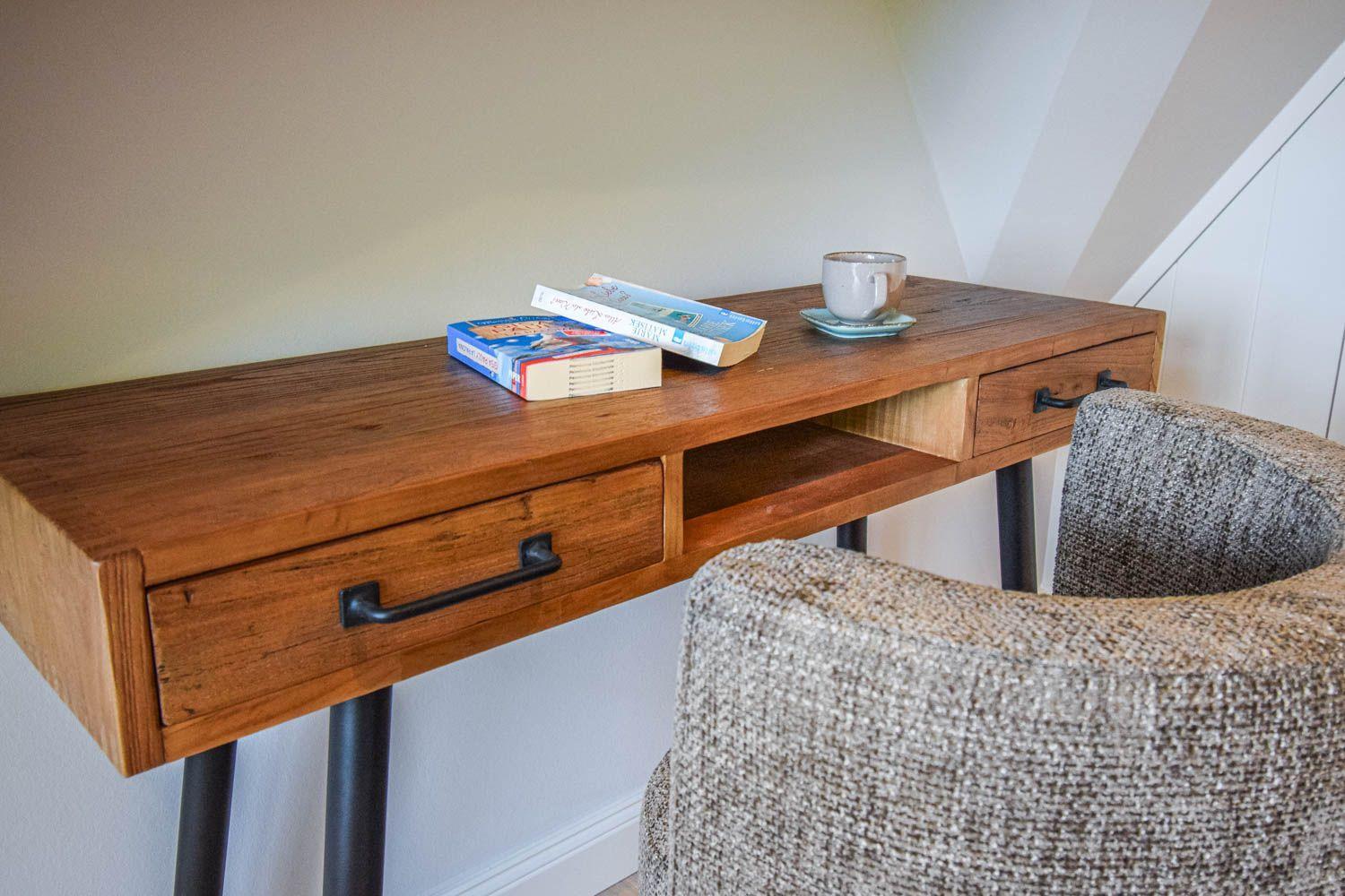 Holzschreibtisch mit zwei Schubladen und Stuhl. Darauf liegen Bücher und eine Tasse.