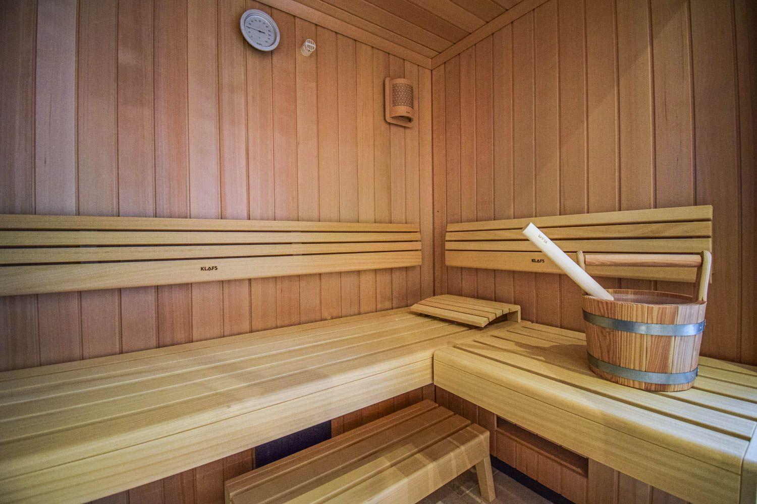 Sauna mit Holzbank, Eimer und Besen. Wandthermometer und Lüftungseinheit sichtbar.
