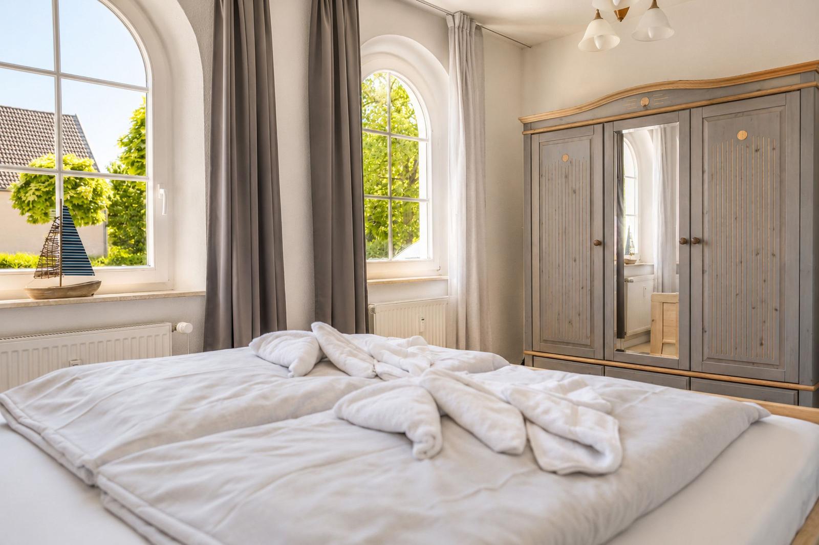 Schlafzimmer mit Bett, Kleiderschrank und zwei Bogenfenstern mit Blick auf Grün.