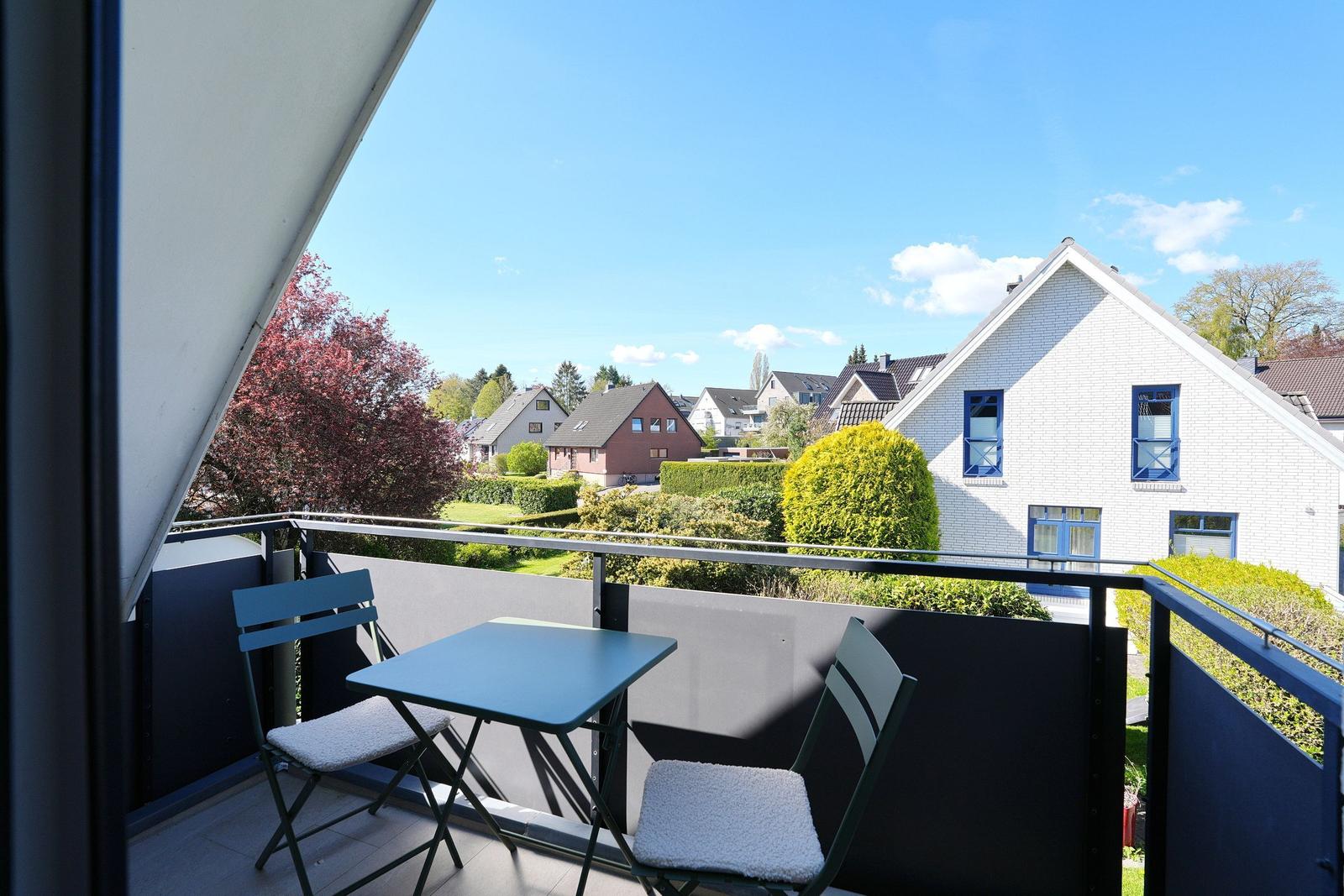 Balkon mit Tisch und zwei Stühlen, Blick auf Wohnhäuser und Bäume.