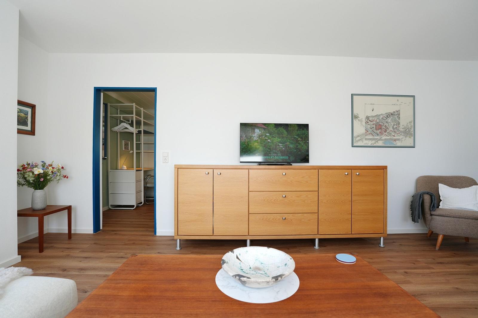 Wohnzimmer mit Holzschrank, Fernseher und Sessel. Durchgang zu Schlafzimmer.