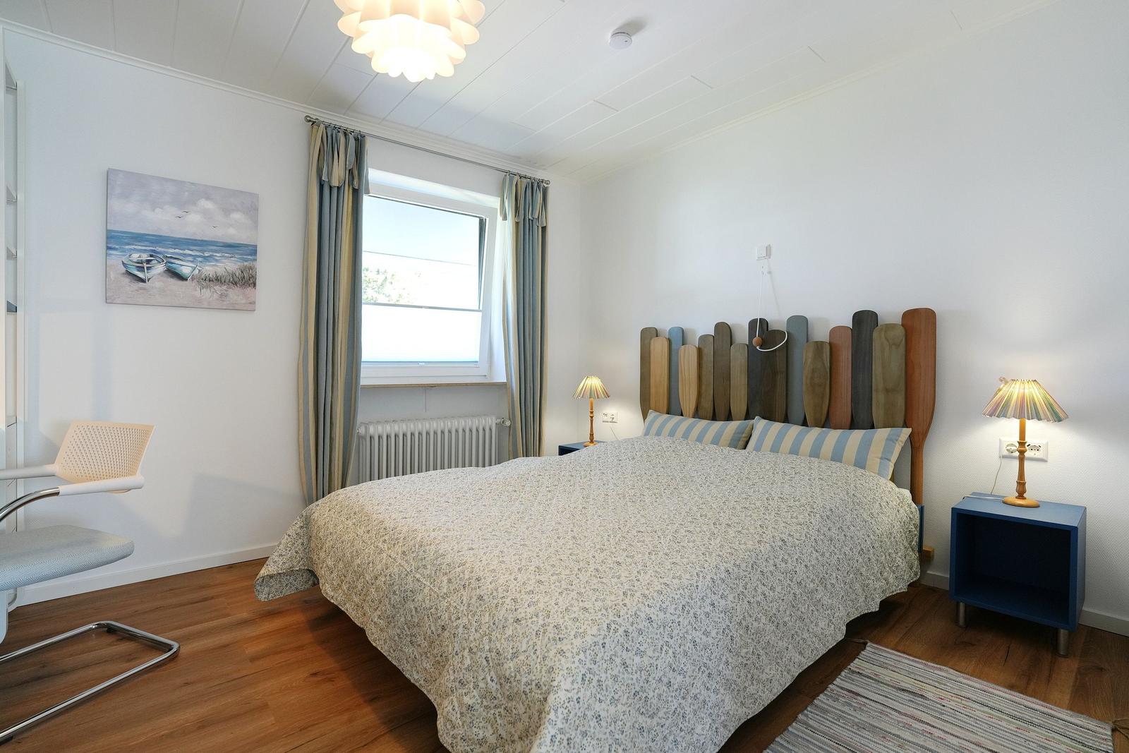 Schlafzimmer mit Bett, Fenster, Vorhängen und Holzkopfteil. Holzfußboden und Wandbild.