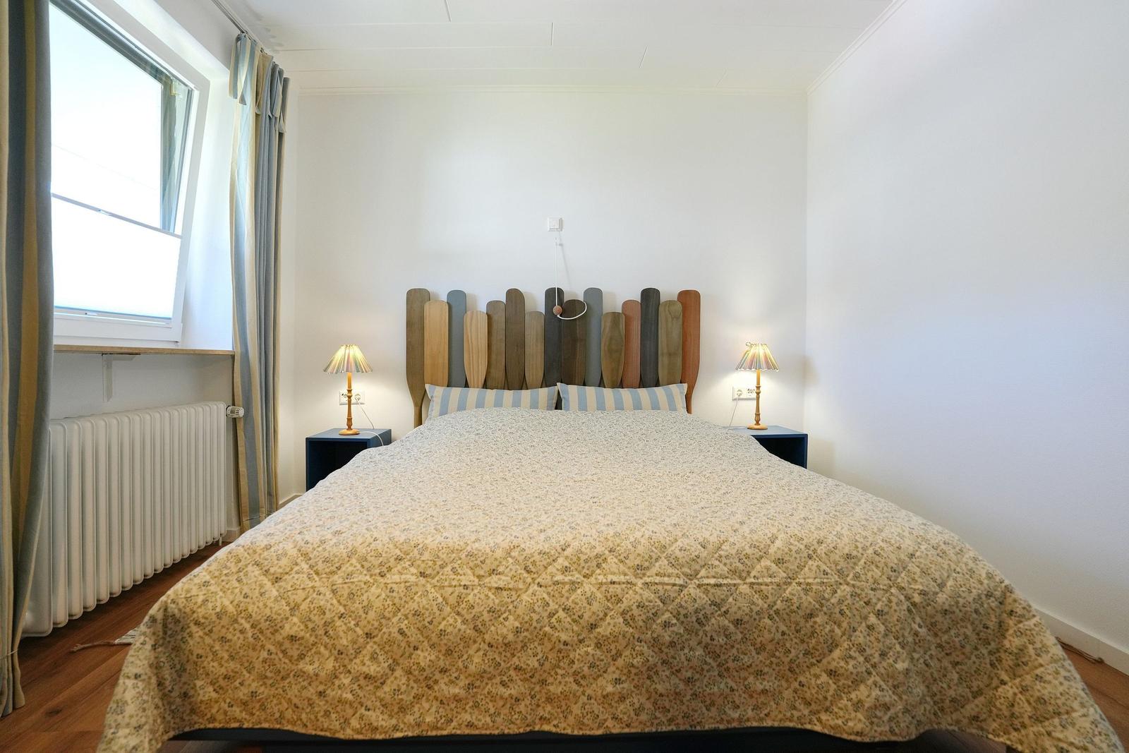 Schlafzimmer mit Bett, Holzkopfteil und Fenster mit Vorhängen.