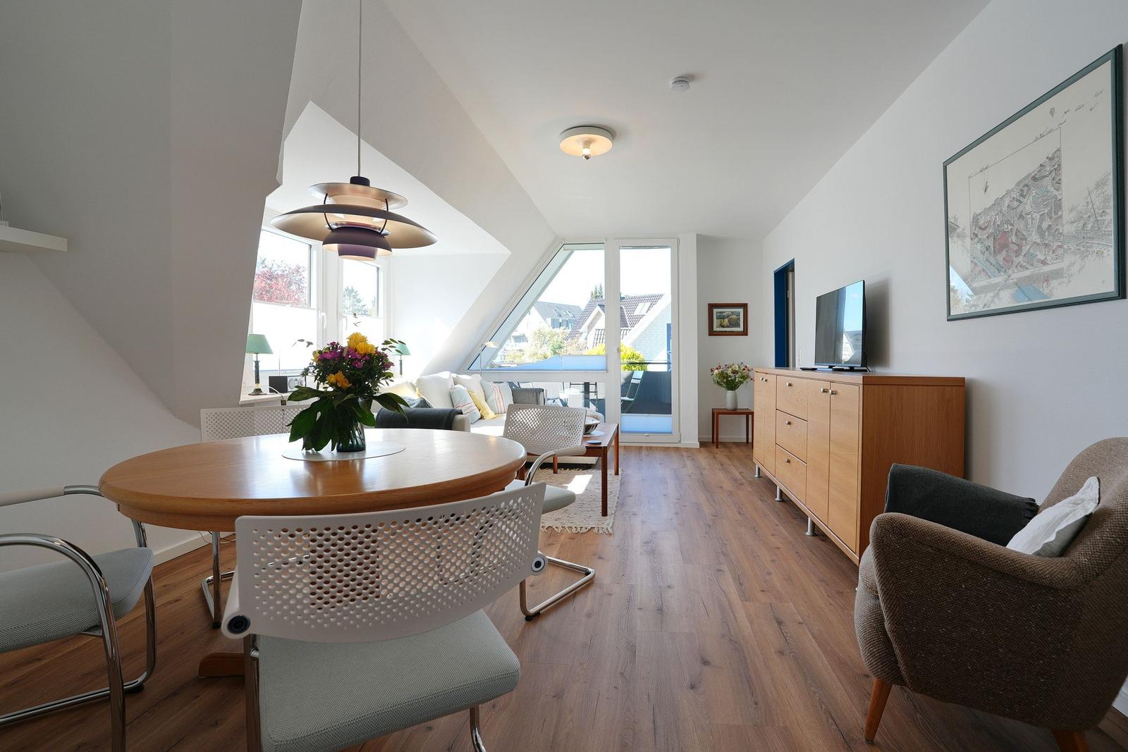 Wohn- und Essbereich mit Holzboden, TV-Schrank und Dachfenster. Tisch mit Stühlen und Blumen.