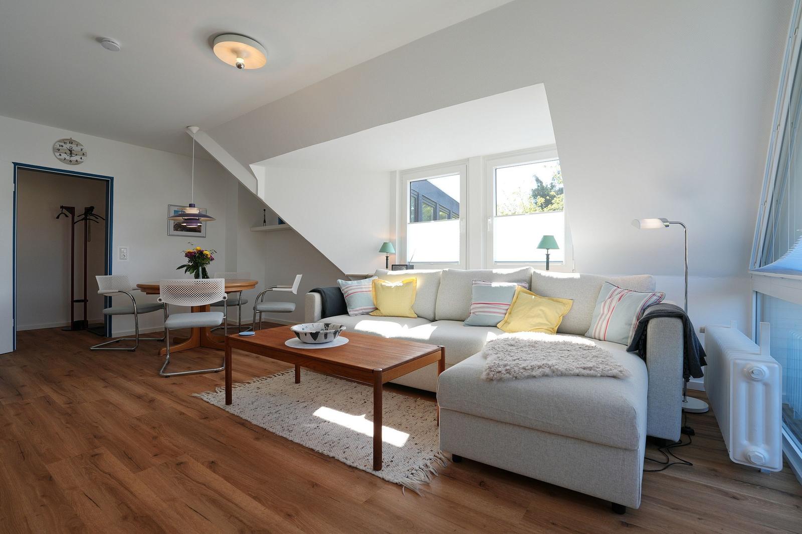 Wohn- und Essbereich mit Sofa, Tisch und Stühlen. Holzboden und große Fenster.