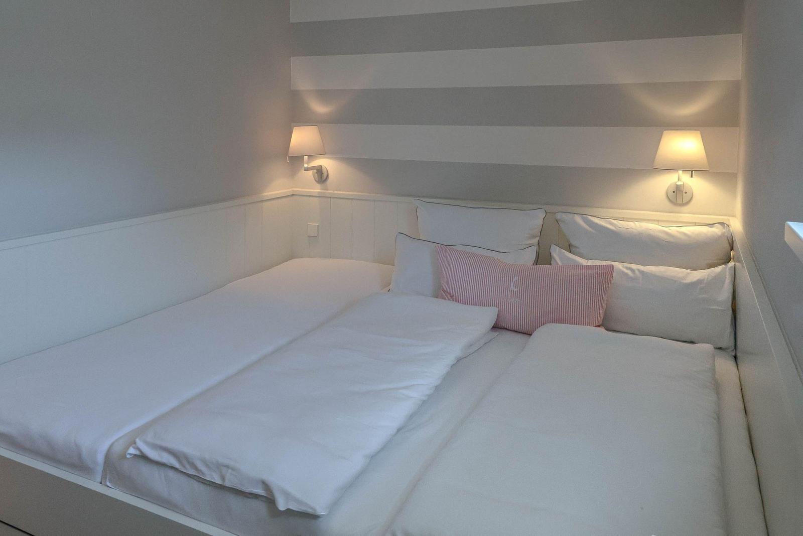 Doppelbett mit weißen Laken und einem rosa Kissen. An der Wand hängen zwei Lampen.
