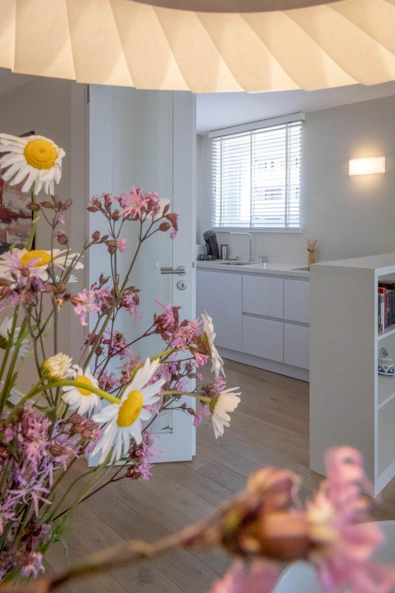 Küche mit weißen Schränken und Fenster mit Jalousie. Vordergrund: Blumenstrauß mit Kamillen und rosa Blüten.