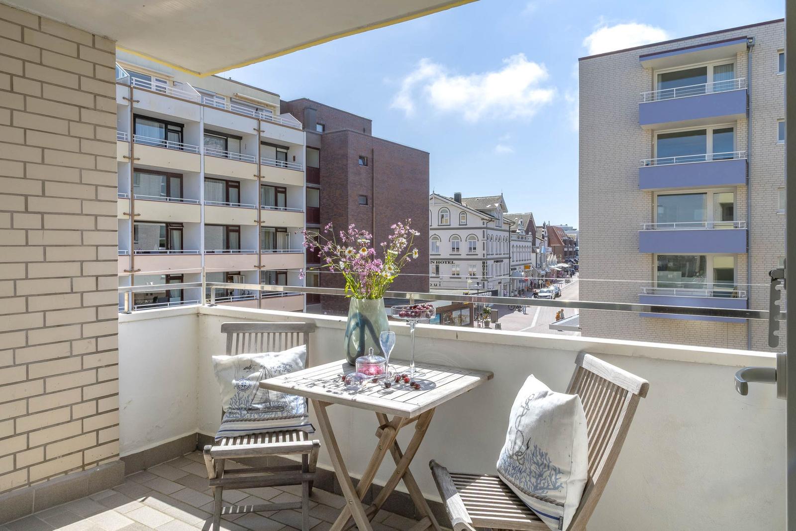 Balkon mit Tisch, zwei Stühlen und Blumen. Blick auf Straße und Gebäude.