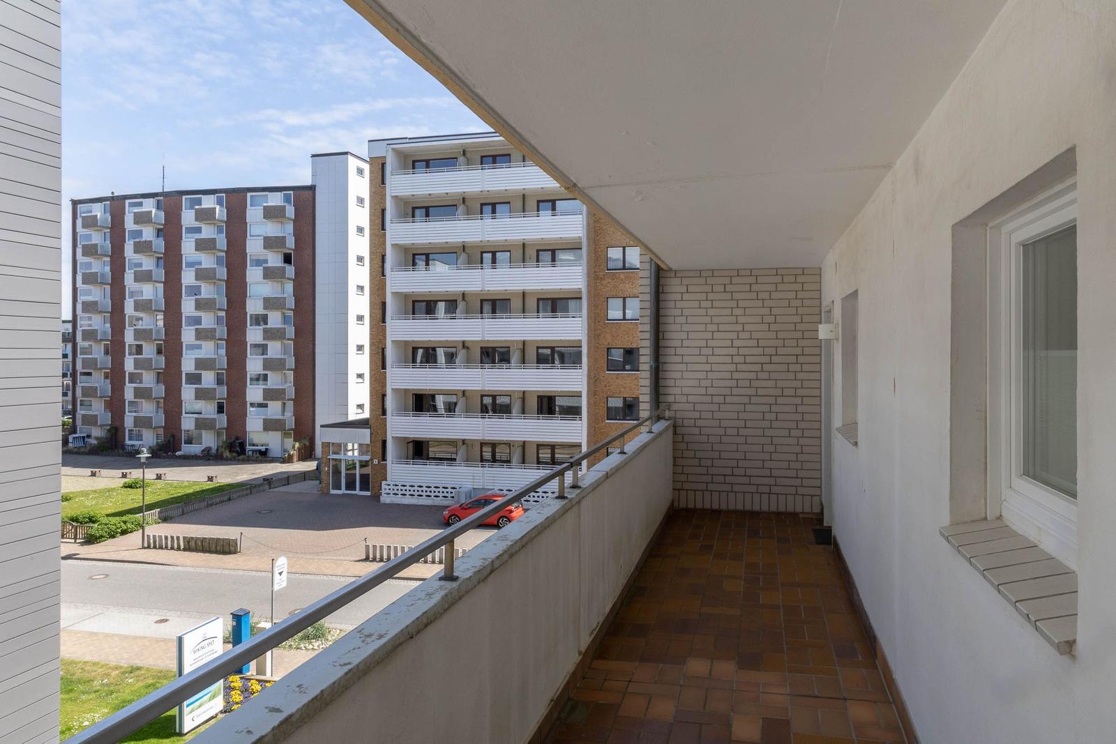 Balcony with tiled floor and railing, overlooking apartment buildings and a red car.