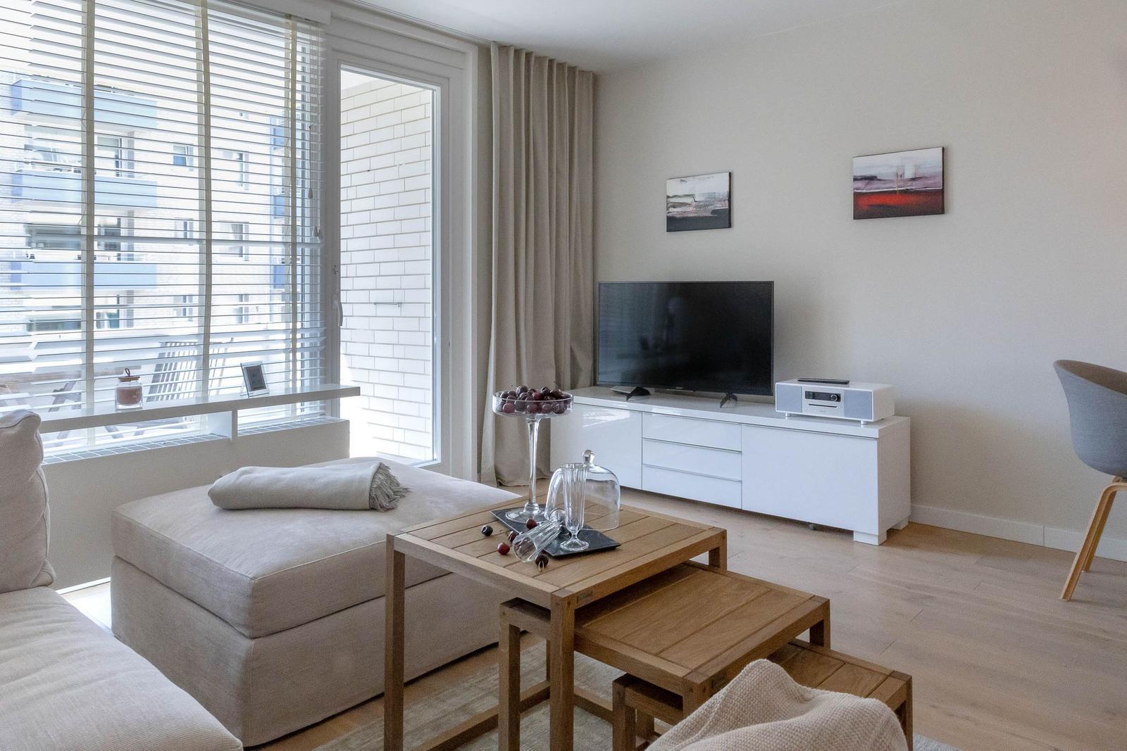 Wohnzimmer mit Sofa, TV-Schrank und Holztisch. Fenster mit Jalousien und Blick auf Balkon.