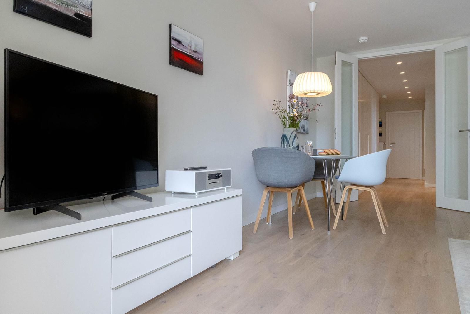 Wohn- und Essbereich mit TV, Tisch und Stühlen. Holzboden und helle Wände.