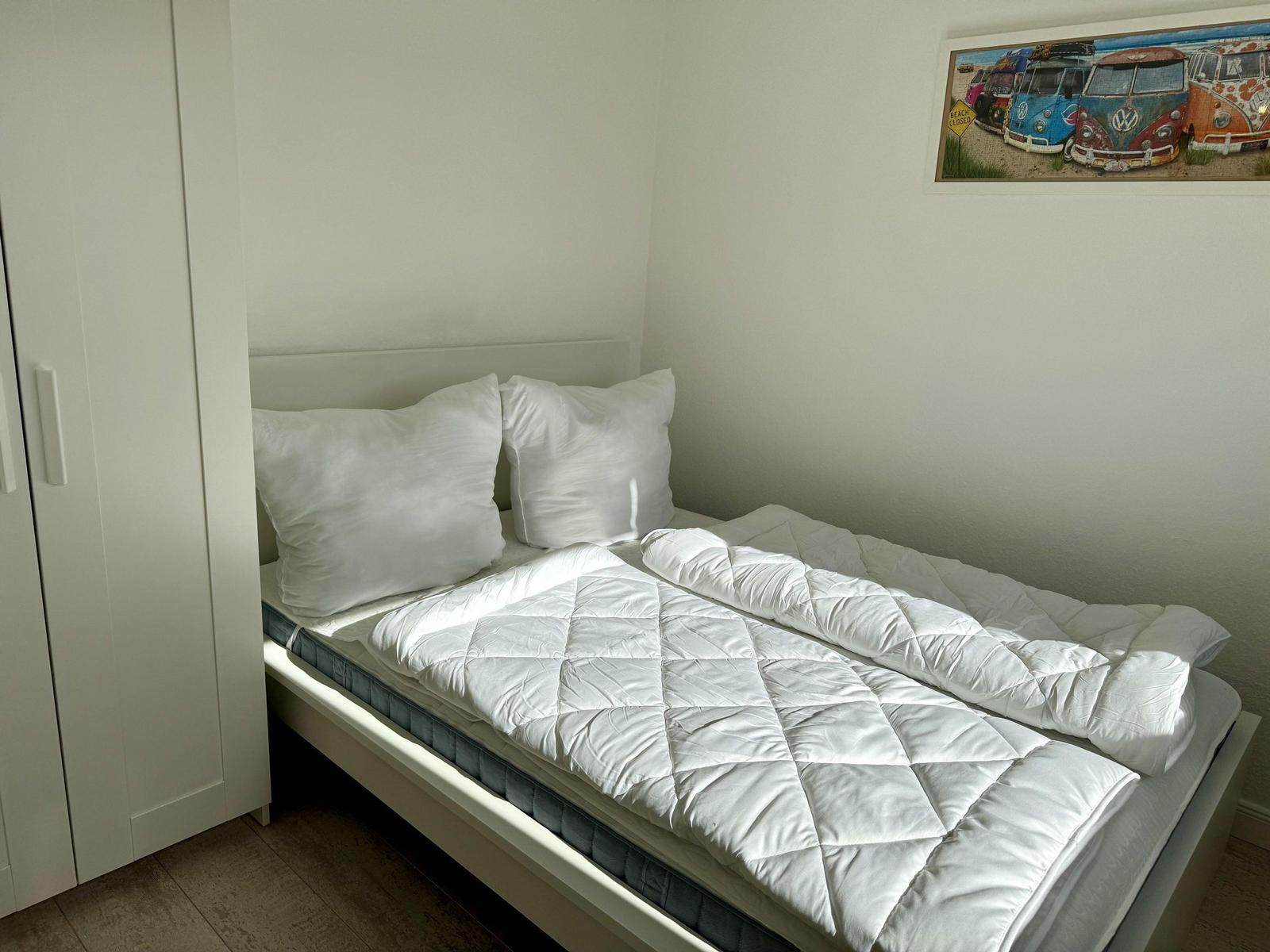Ein Bett mit weißer Bettwäsche und Kissen. An der Wand hängt ein Bild von VW-Bussen.