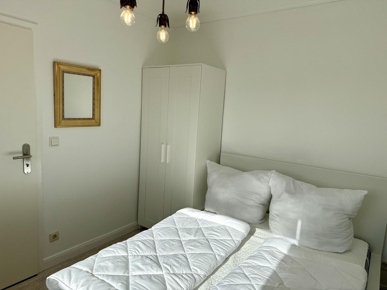Schlafzimmer mit weißem Bett, Kleiderschrank und Spiegel an der Wand.
