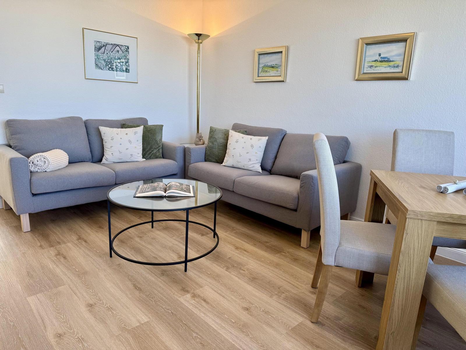 Zwei graue Sofas, runder Tisch, Holzfußboden und Wandbilder.